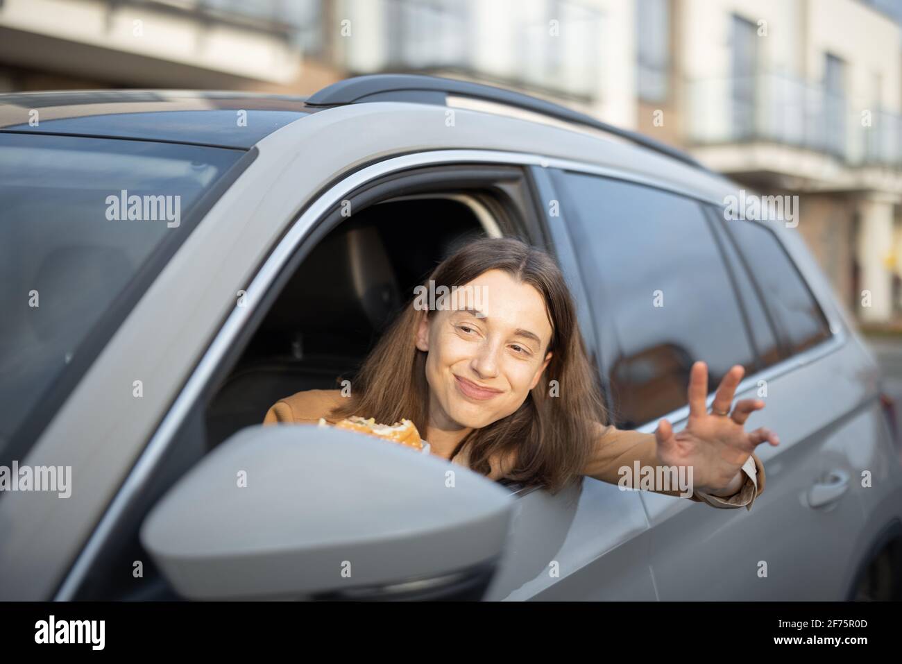 Happy woman eating a burger in the car while greet someone on the road. Have unhealthy fast food snack. Food to go. Hungry and busy concept. Stock Photo