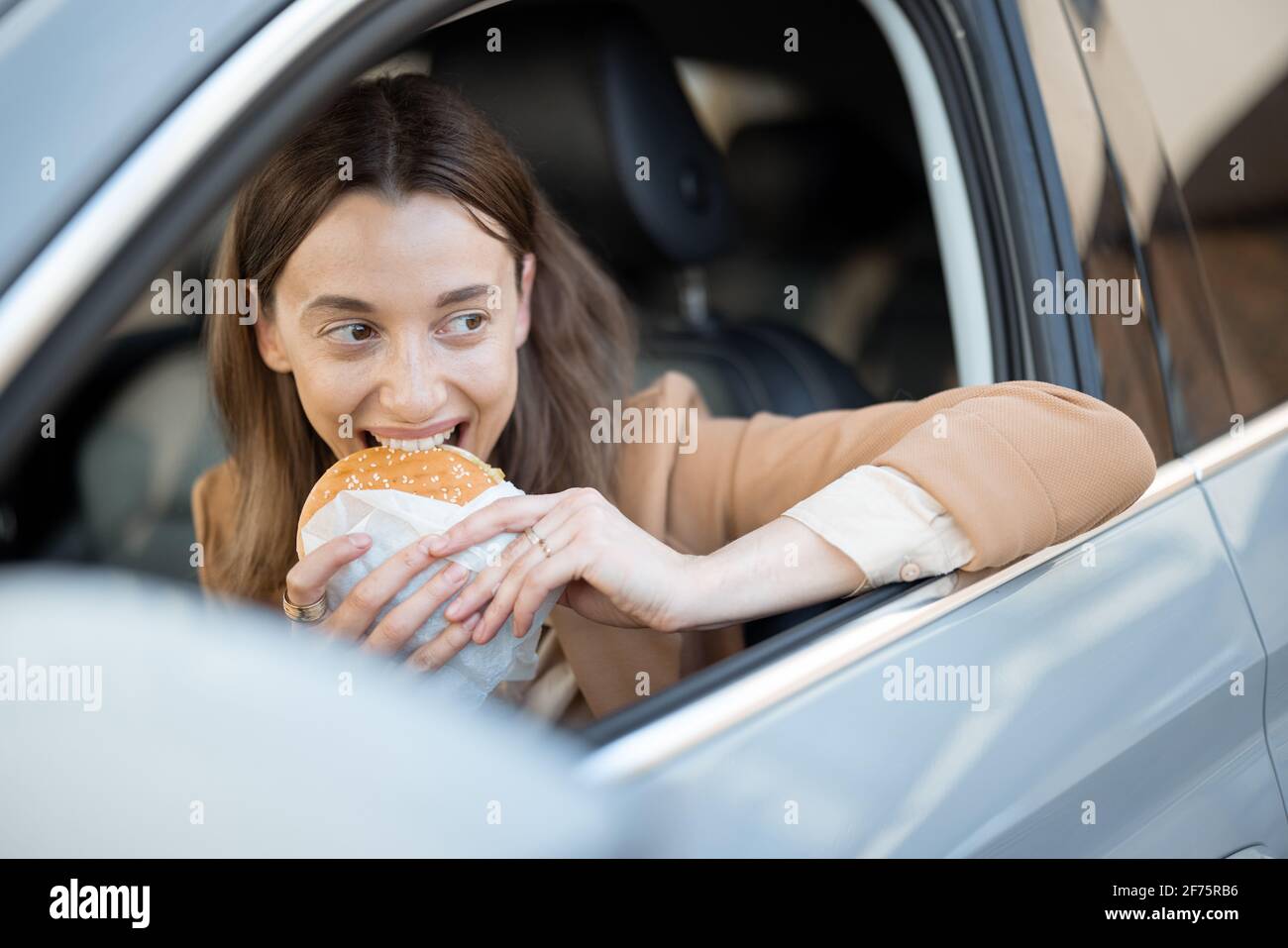 Happy woman eating a burger in the car. Bites a sandwich. Have unhealthy fast food snack. Food to go. Hungry and busy concept. Stock Photo