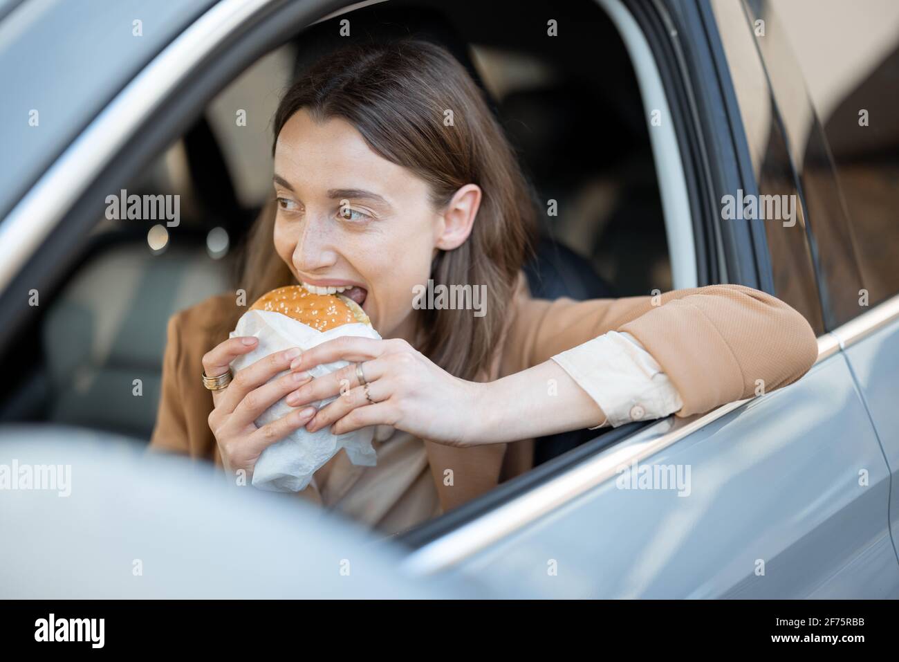 Happy woman eating a burger in the car. Bites a sandwich. Have unhealthy fast food snack. Food to go. Hungry and busy concept. Stock Photo