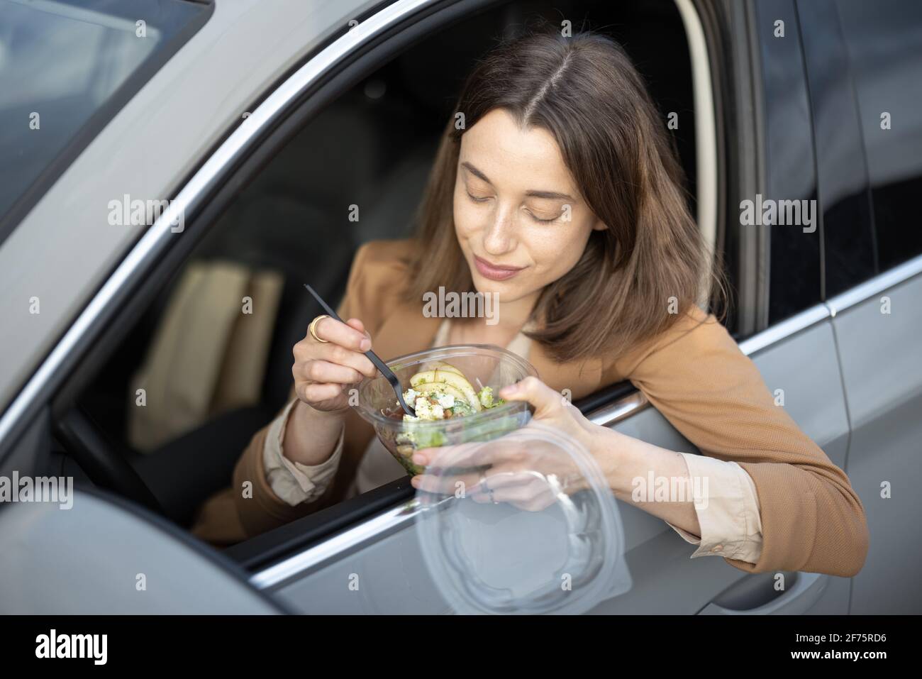 Beautiful woman eating heathy salad in the car. Received a food order to go. Have a quickly snack on the Lunch Break.  Stock Photo