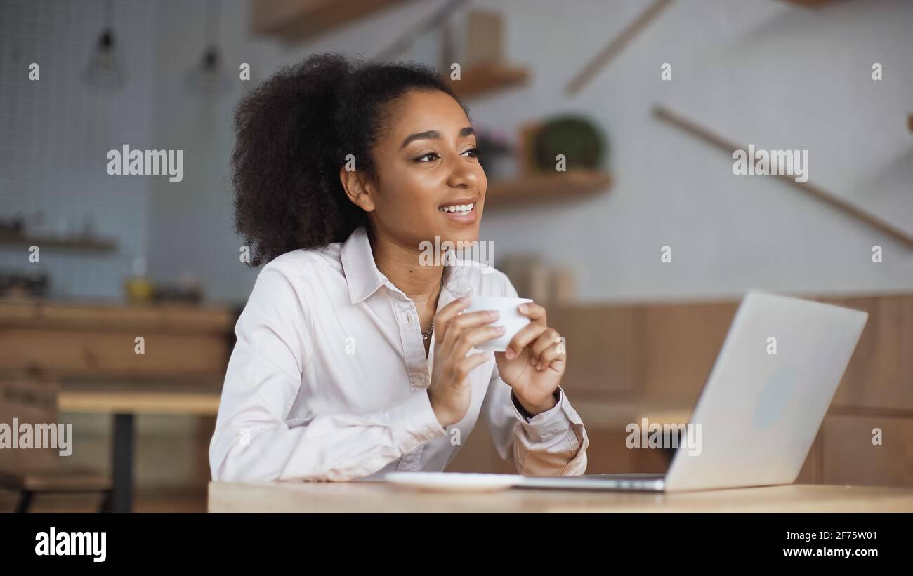 happy african american woman holding cup of coffee near laptop in cafe ...