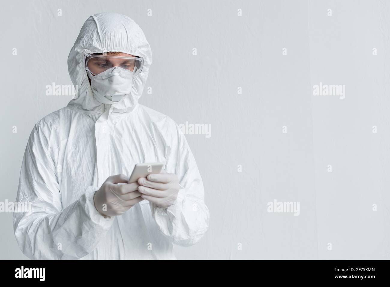 Scientist in hazmat suit using smartphone near wall Stock Photo