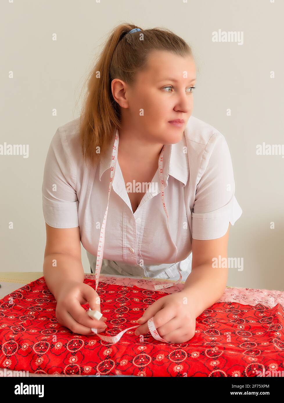 The woman is leaning over the table on which the cloth is lying, with a thoughtful look, holding a crayon and a tailor's meter Stock Photo