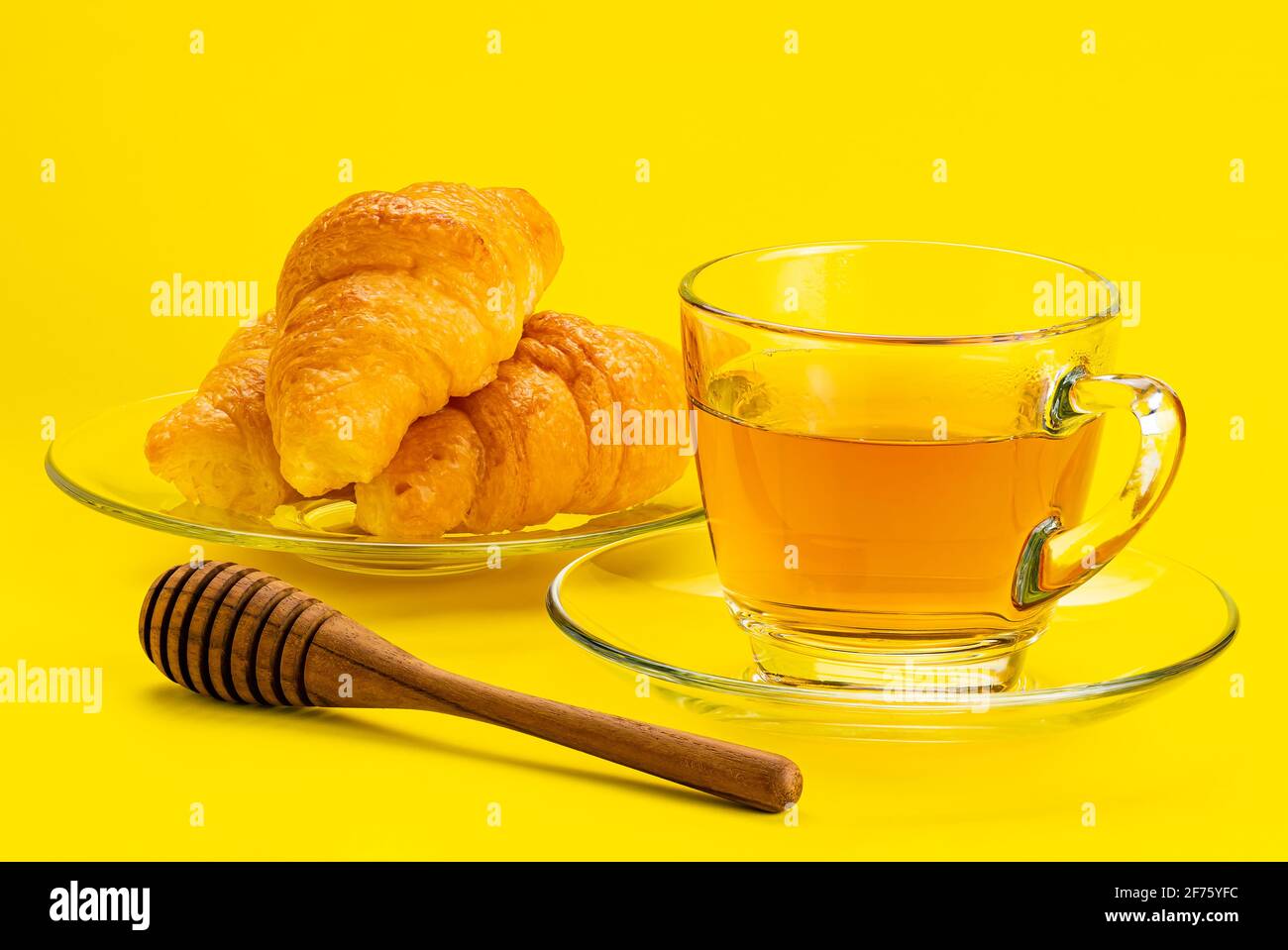 Freshly prepare croissant in a glass plate a cup of tea and a wooden honey dipper on yellow background. Stock Photo