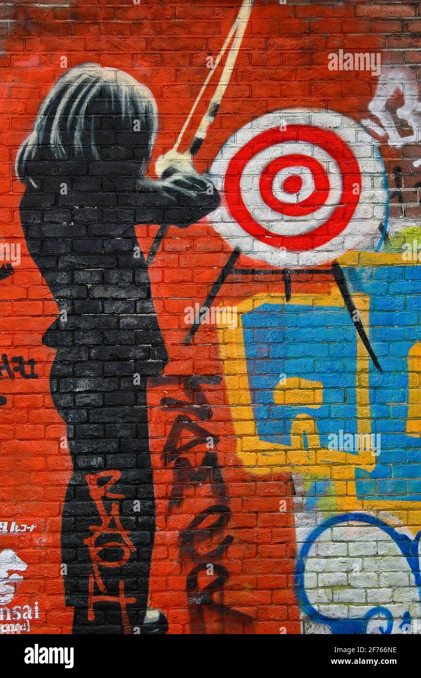 Taking aim … a female archer wearing a dark suit prepares to fire an arrow at a target on a brick wall sprayed bright red in psychedelic street art on one of the former long-term squatters’ strongholds close to the world-famous Slangenhuis or Snake House in Spuistraat, Amsterdam, North Holland, Netherlands. Stock Photo