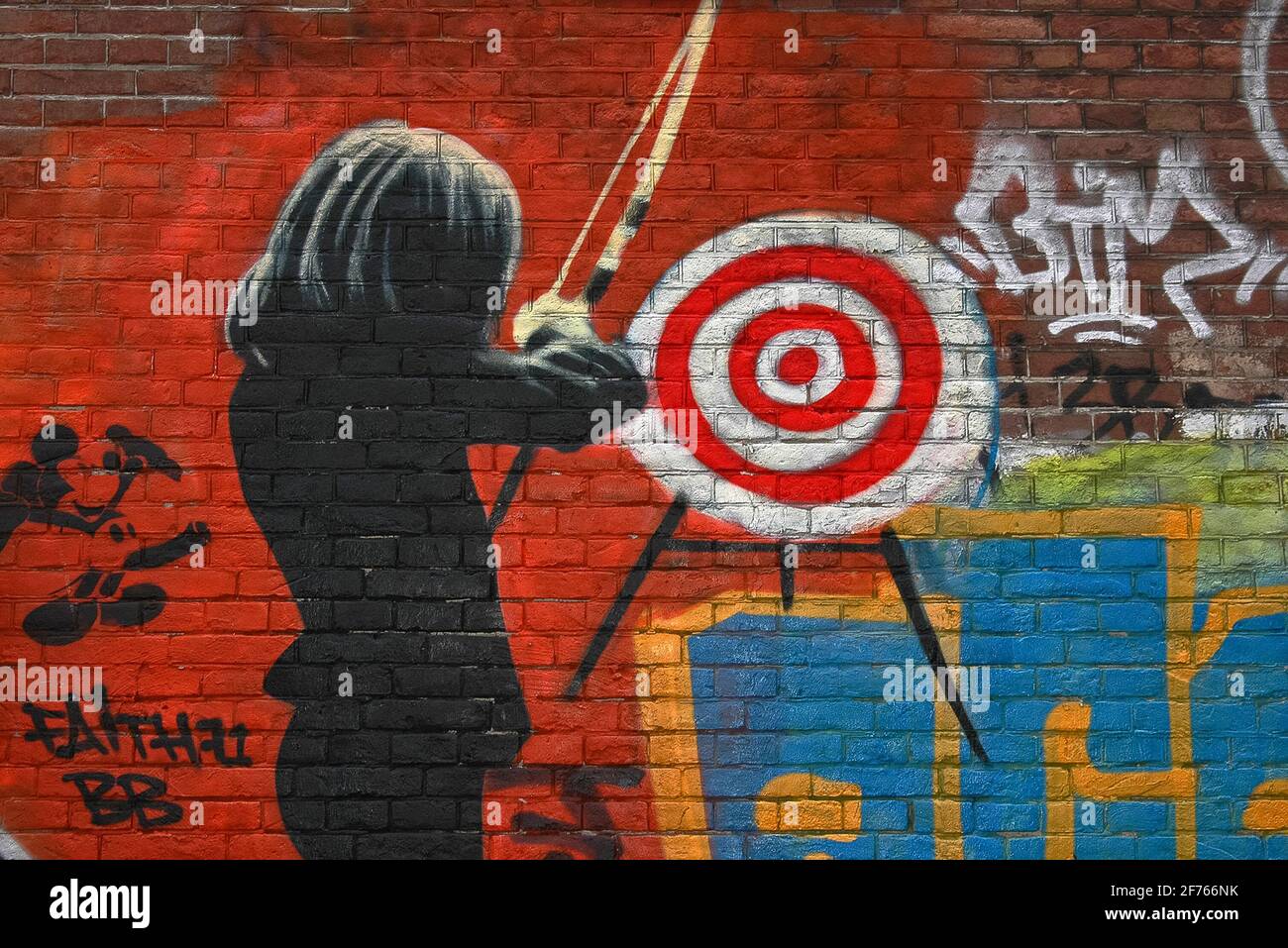 A female archer in a dark suit prepares to fire an arrow at a target on a brick wall sprayed bright red in psychedelic street art on one of the former long-term squatters’ strongholds close to the world-famous Slangenhuis or Snake House in Spuistraat, Amsterdam, North Holland, Netherlands. Stock Photo