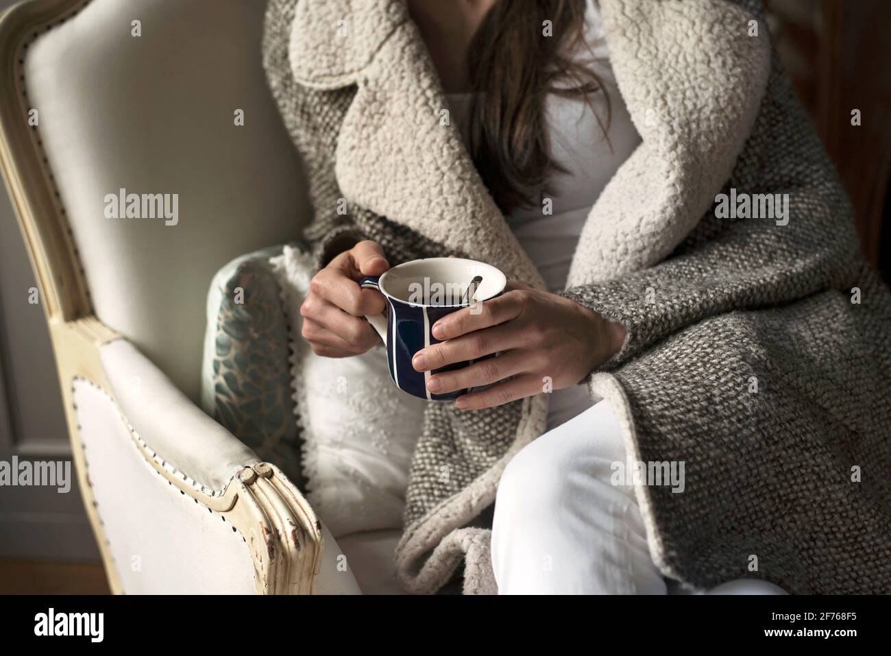 Close up of female in cosy warm coat sitting on vintage armchair holding a cup of coffee. Indoor lifestyle with relaxed country vibes. London, UK Stock Photo