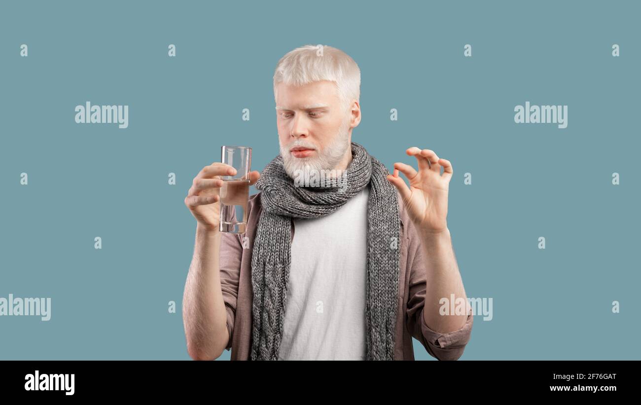 Illness treatment. Sick albino man holding pill and glass of water, taking medicine over turquoise background Stock Photo