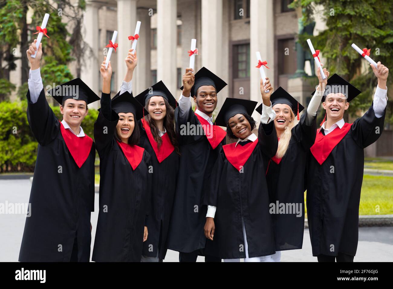 Cheerful multiracial students in graduation costumes raising diplomas ...
