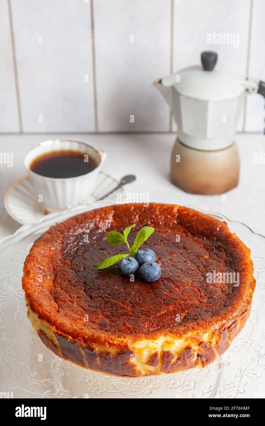Homemade Basque burnt cheesecake on a plate with blueberries and mint leaves on light background,with cup of coffee and geyser coffee maker. Stock Photo