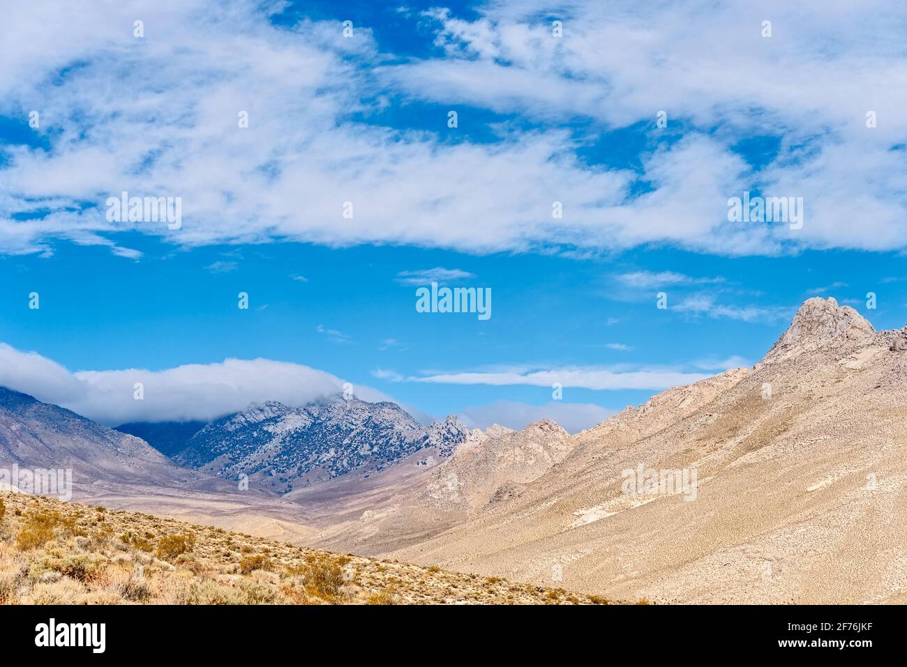 Blue mountains with white clouds following the contour of the peak. Barren brown mountain peak under blue skies with white fluffy clouds. Stock Photo