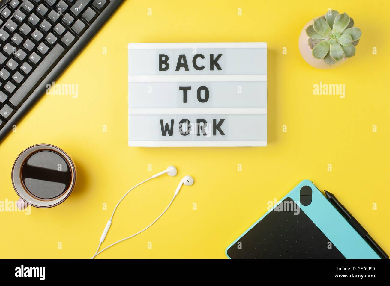 Back to work - text on display lightbox on yellow background workplace. Black keyboard, white earphones, coffee, tablet. Stock Photo
