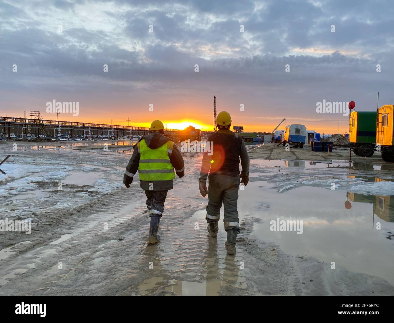 Two oil workers in work clothes walk through the site along a muddy road to an oil rig in the background of the sunset. Heavy male work of drillers. Stock Photo