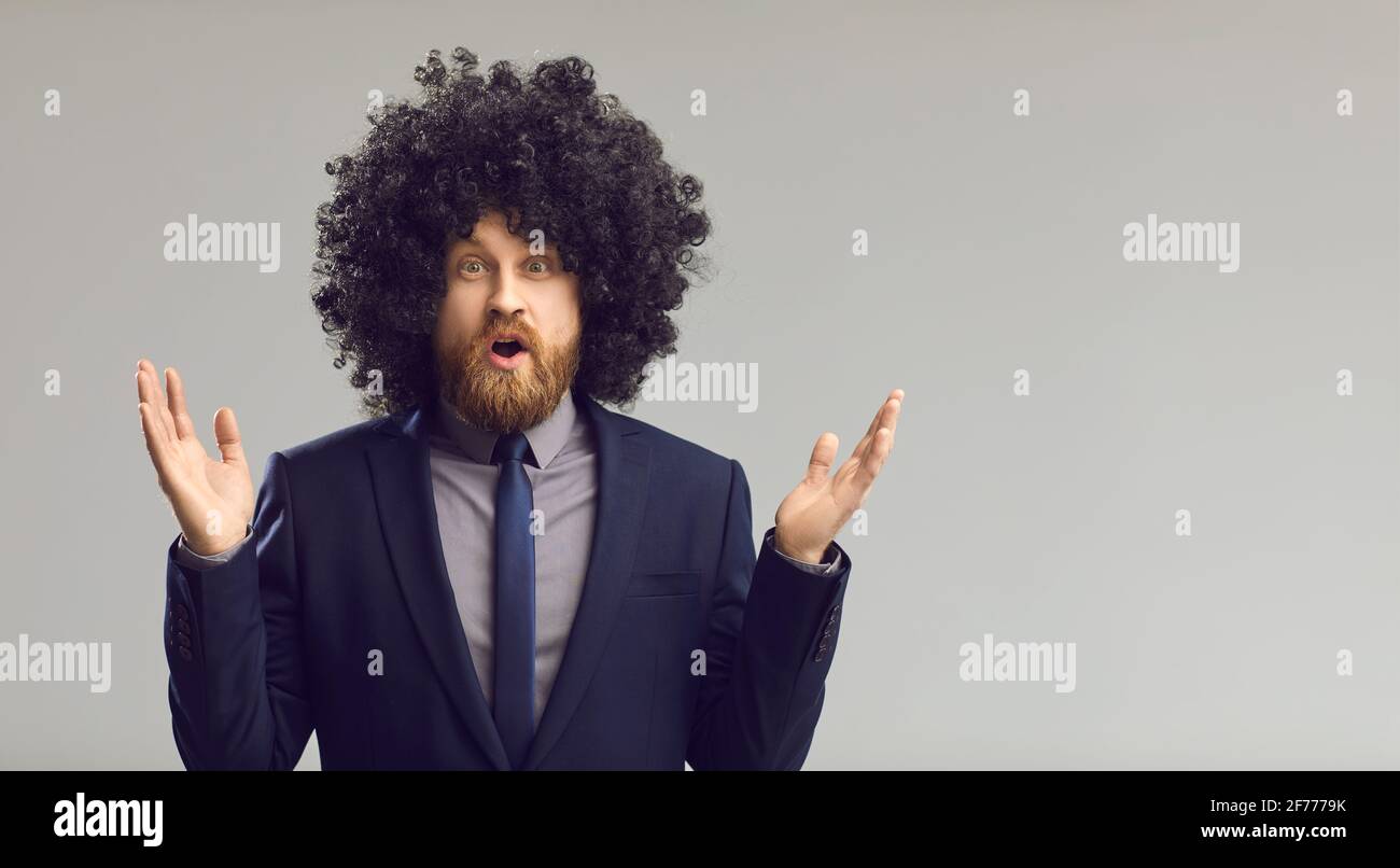 Funny young man in curly wig gasps in surprise standing on gray copy space background Stock Photo