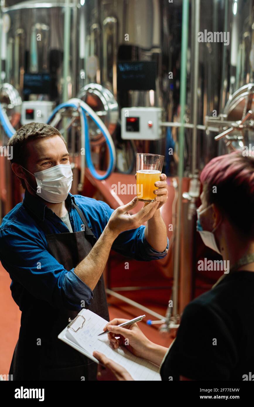 Young male brewer in protective mask holding glass of fresh beer after preparation and discussing its characterstics with female colleague Stock Photo