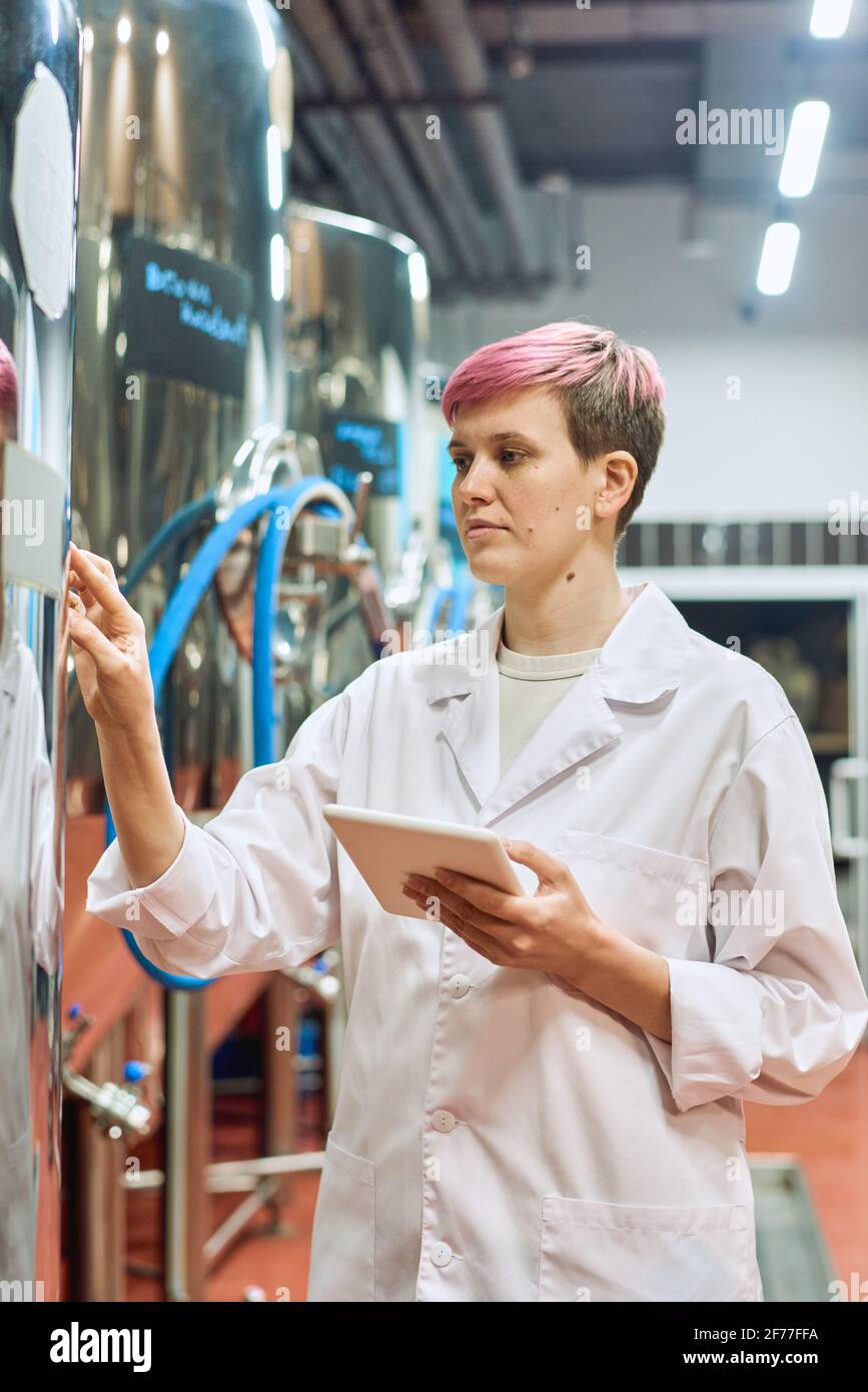 Young confident female expert in whitecoat using tablet while touching control panel of beer production equipment in workshop Stock Photo