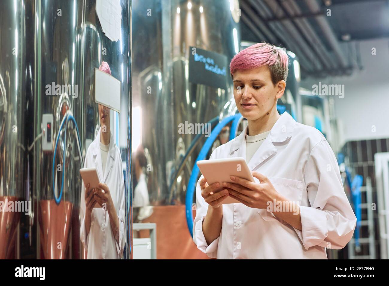Young female brewery expert in whitecoat looking through data on screen