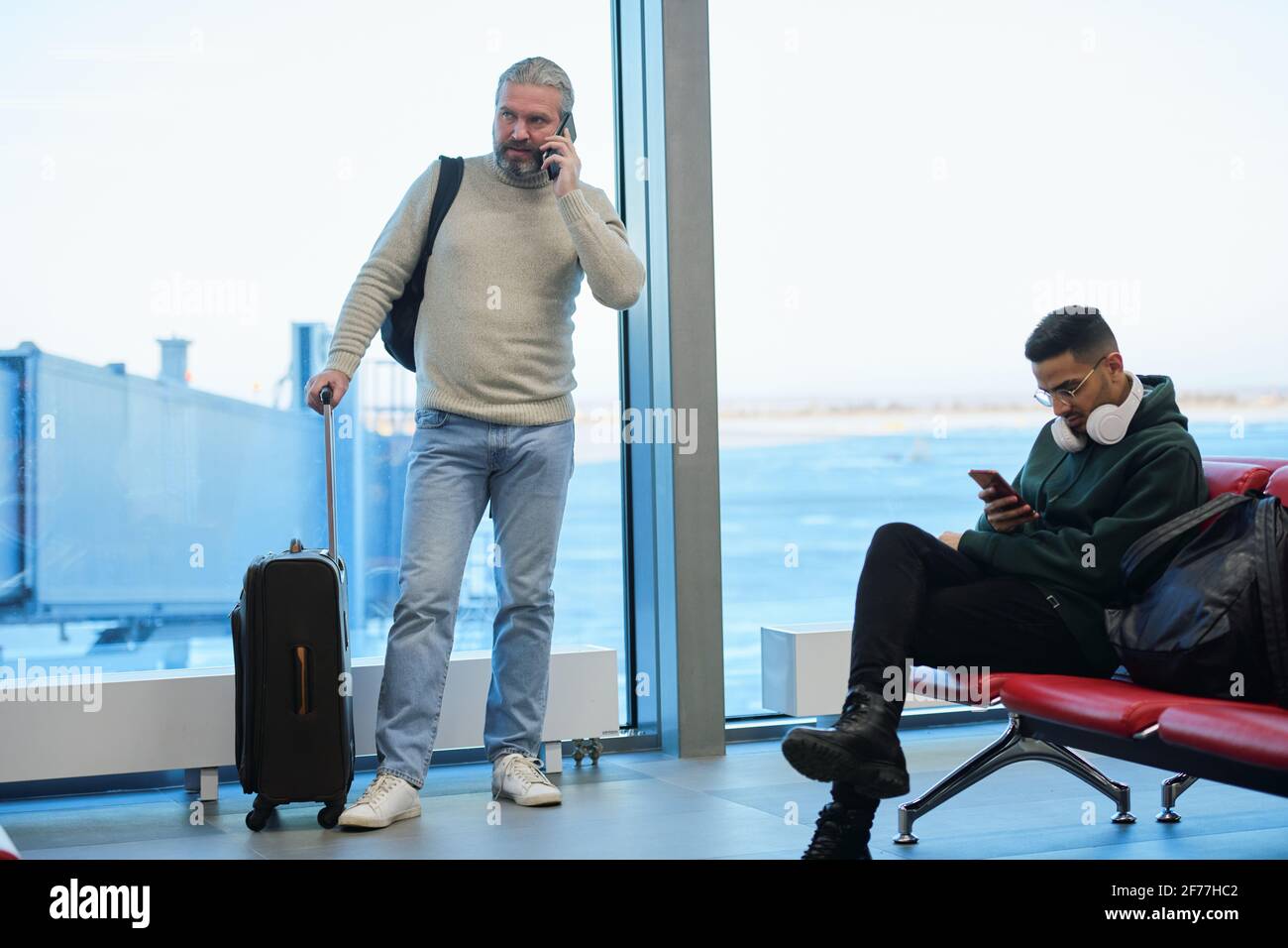 Mature man using smartphone during telephone conversation while young male traveler scrolling through contacts or posts in airport lounge Stock Photo