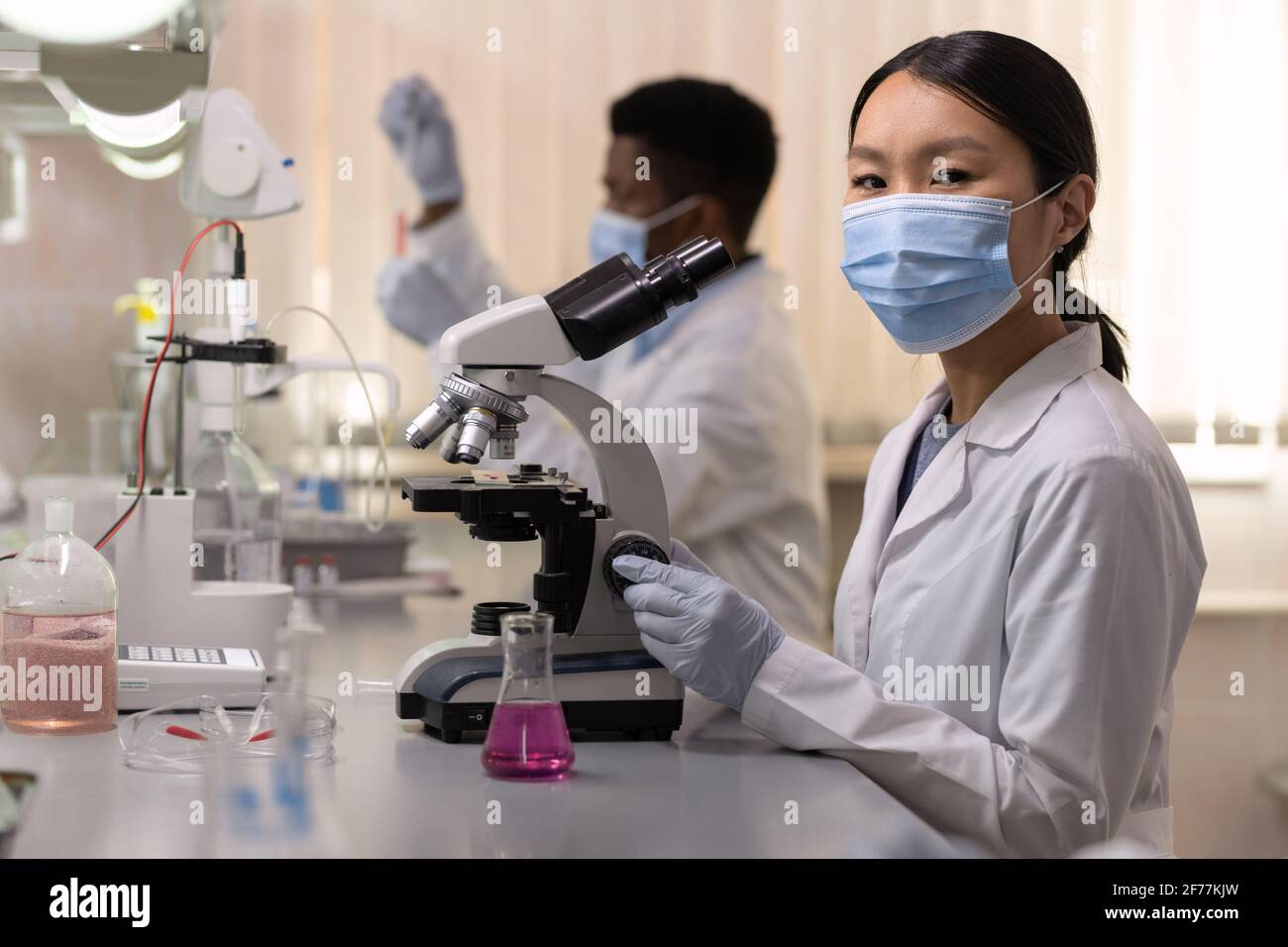 Portrait of Asian young scientist in protective mask looking at camera while working with microscope at the table in the lab Stock Photo