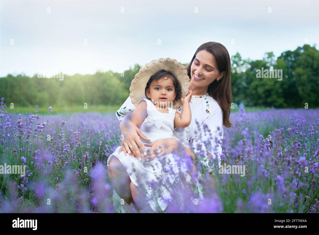 Young happy mother sitting between purple flowers with kid wearing straw hat and looking at camera. Smiling woman posing with baby daughter on knees in lavender field. Concept of nature, motherhood. Stock Photo