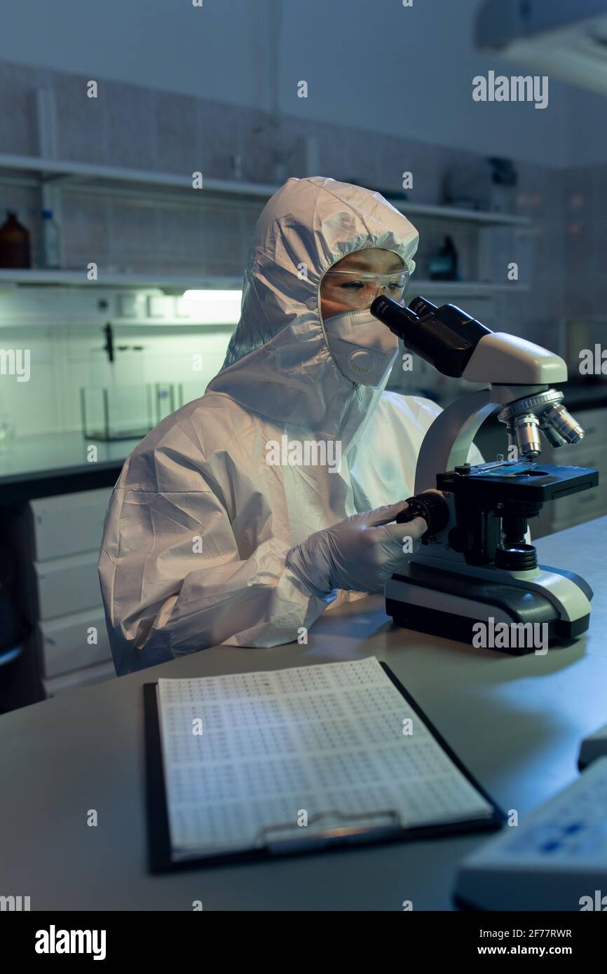 Female chemist in protective wear sitting at her workplace and looking through the microscope while working in the lab Stock Photo