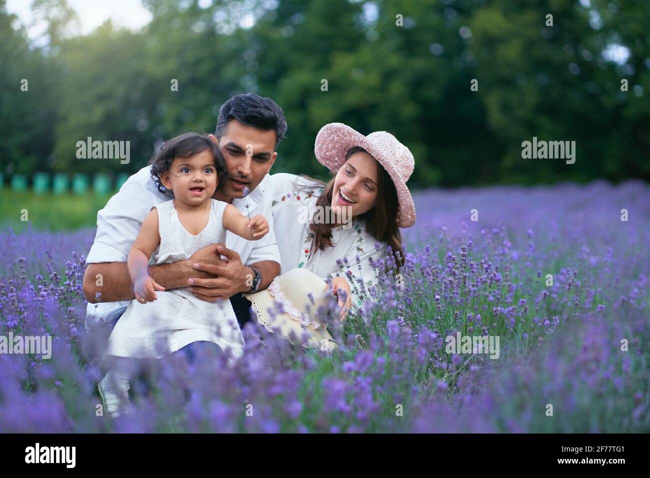 Front view of young mother, father and daughter posing in lavender meadow and smiling. Adorable baby girl holding flower and enjoying time with loving parents outdoors. Young family, nature concept. Stock Photo