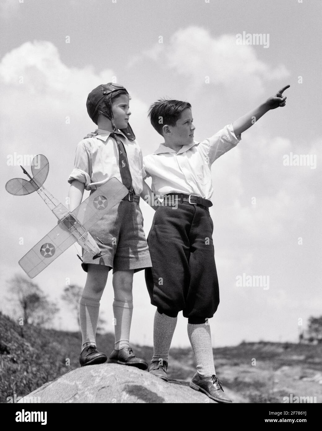 1920s 1930s ONE BOY POINTS TO THE SKY HIS FRIEND A SECOND BOY IS HOLDING A MODEL AIRPLANE AND WEARS AN AVIATOR HELMET - b11921 HAR001 HARS KNICKERS JOY PLANES POINT BROTHERS FLIGHT RURAL HOME LIFE COPY SPACE FRIENDSHIP FULL-LENGTH INSPIRATION MALES PLANNING SIBLINGS B&W GOALS DREAMS ADVENTURE AIRPLANES DISCOVERY AND DREAMING EXCITEMENT LOW ANGLE RECREATION AVIATION NECKTIE SIBLING KNEE SOCKS WEARS AVIATOR IMAGINATION POINTS SUPPORT KNICKERBOCKERS ANOTHER GROWTH HOLDS IDEAS JUVENILES TOGETHERNESS BLACK AND WHITE CAUCASIAN ETHNICITY HAR001 OLD FASHIONED Stock Photo