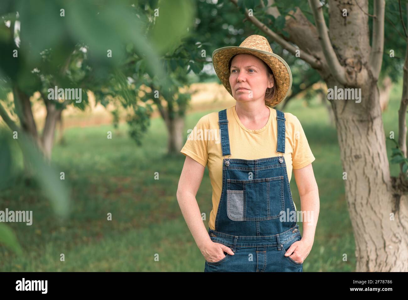 Portrait of female farmer posing in walnut fruit orchard, wearing straw hat and bib overalls Stock Photo