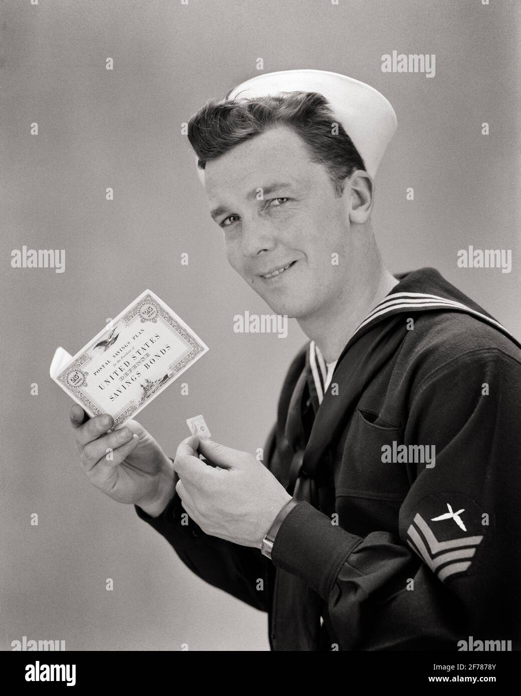 1940s SMILING NAVY SAILOR LOOKING AT CAMERA HOLDING WORLD WAR 2 UNITED STATES SAVINGS BOND POSTAL PLAN BOOKLET AND STAMP  - d1634 HAR001 HARS STUDIO SHOT NAVY UNITED STATES COPY SPACE HALF-LENGTH PERSONS POSTAL UNITED STATES OF AMERICA MALES FORCE B&W BOND NORTH AMERICA EYE CONTACT FREEDOM GOALS NORTH AMERICAN CHEERFUL NAVAL AND WORLD WARS WORLD WAR WORLD WAR TWO WORLD WAR II SMILES UNIFORMS FORCES BOOKLET JOYFUL NAVIES PATRIOTIC WORLD WAR 2 USN YOUNG ADULT MAN BLACK AND WHITE CAUCASIAN ETHNICITY HAR001 OLD FASHIONED Stock Photo