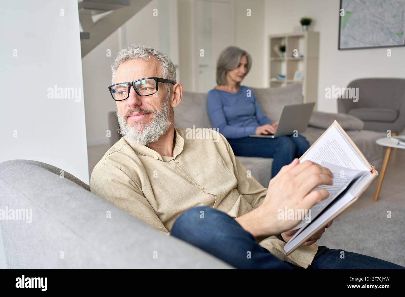 Happy older senior man husband reading book relaxing sitting on couch at home. Stock Photo