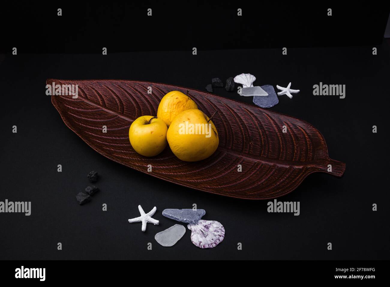 Three yellow apples placed on a brown leaf-shaped plate with a few shells, on a black background Stock Photo