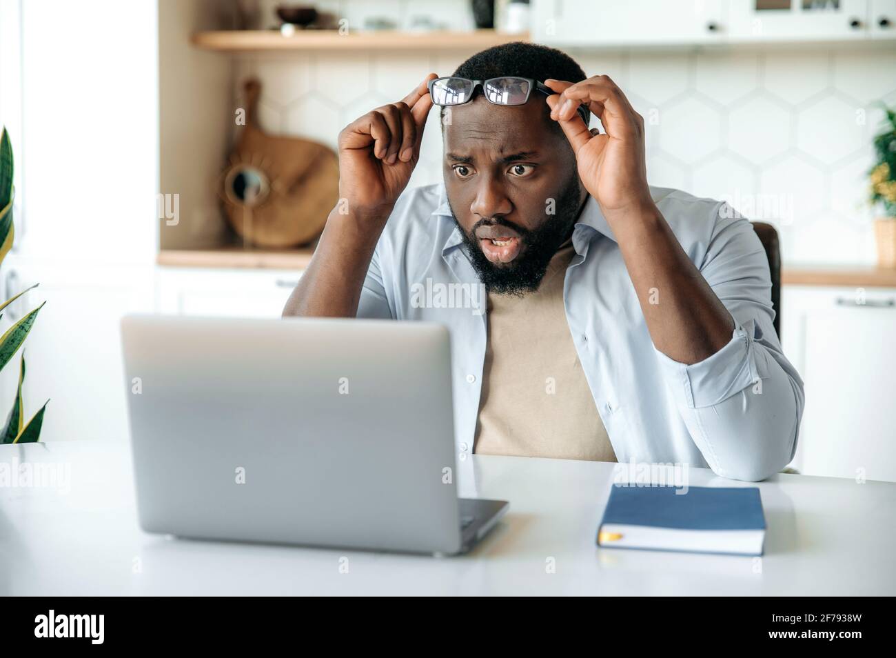 Amazed bewildered African American bearded man, business executive, freelancer sits at his desk, looks at his laptop in surprise while taking off glasses, reads unexpected news or message Stock Photo