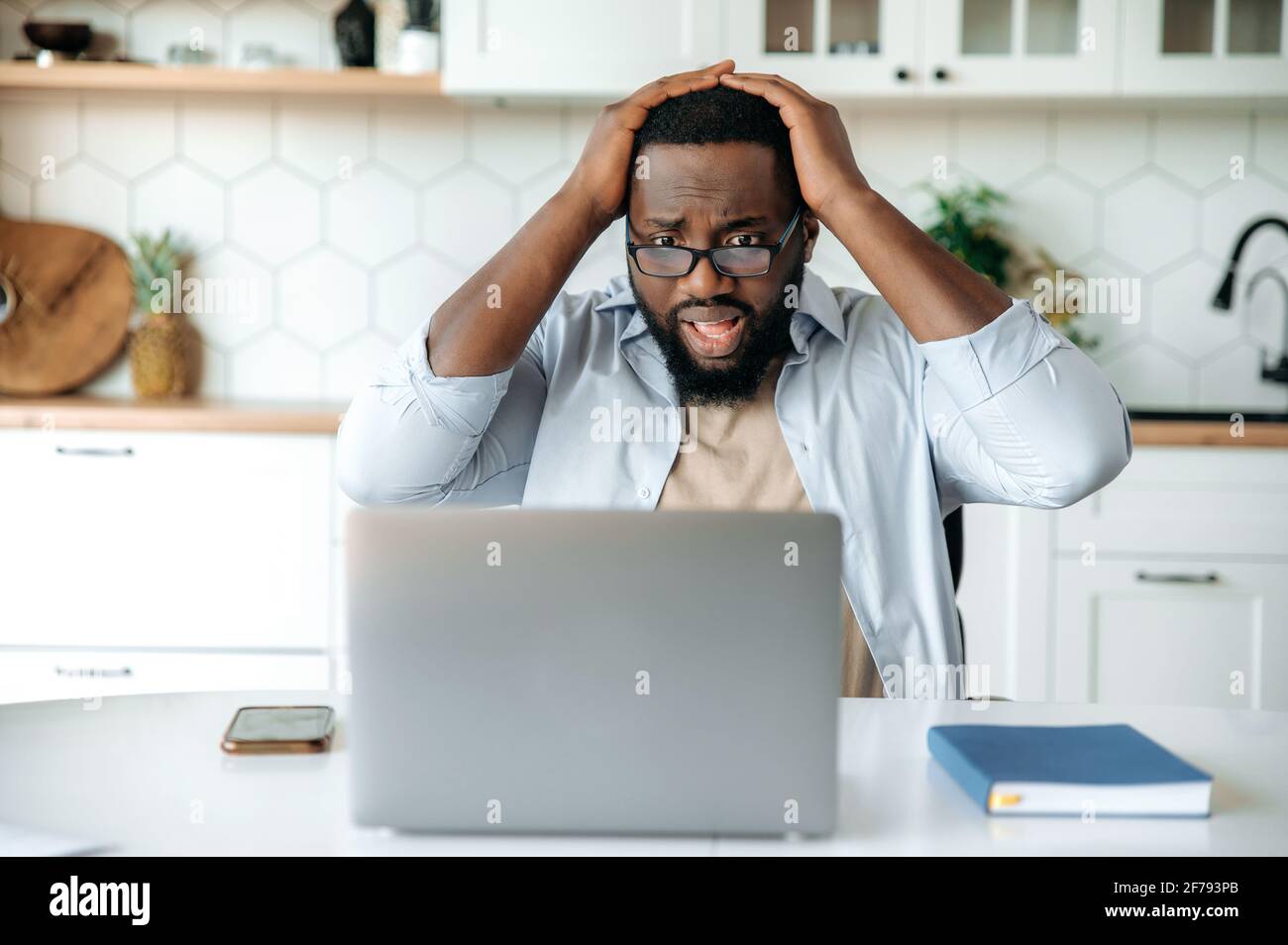 Shocked African American bearded man with glasses sits at his desk at home, looks at his laptop in confusion, reads bad news, received an unexpected message, failed deal, holds head with hands Stock Photo