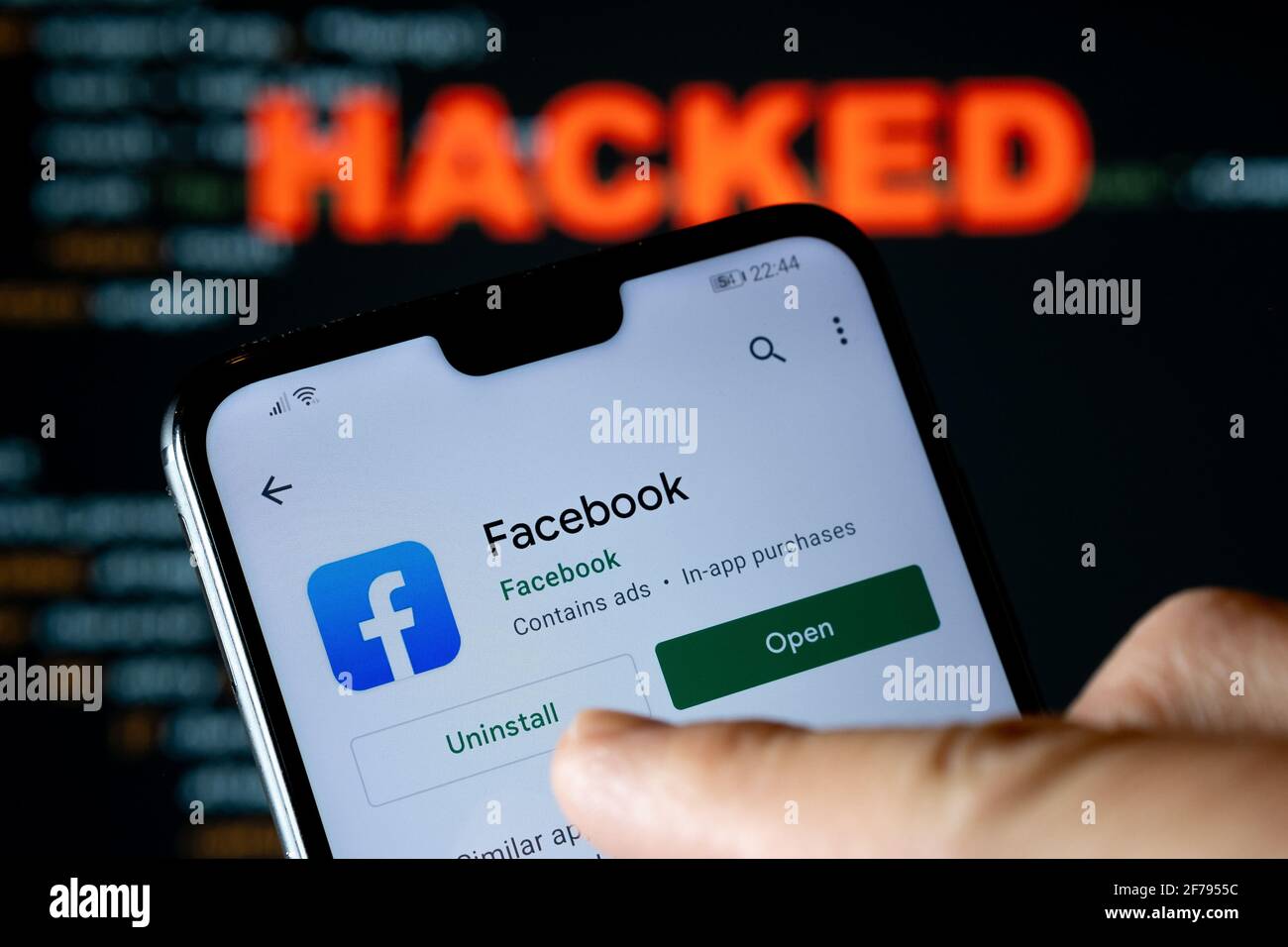 Facebook app in Play market seen on the smartphone, finger pointing at it and blurred HACKED word on the blurred background. Concept. Stafford, United Stock Photo