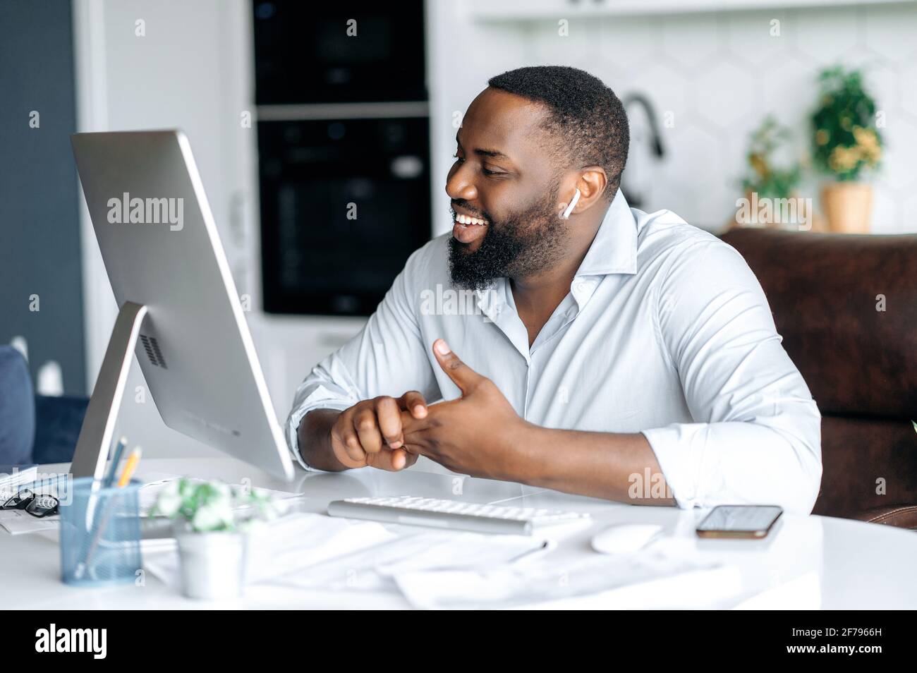 Joyful pleasant successful african american man, lawyer or real estate agent working remotely at computer, talking to colleague or customer via video call uses wireless headset, smiling friendly Stock Photo