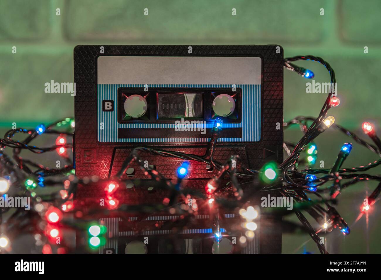 An audio cassette in a black plastic case is reflected on a black matte surface. Colorful lights are garlanded around it. Close-up Stock Photo