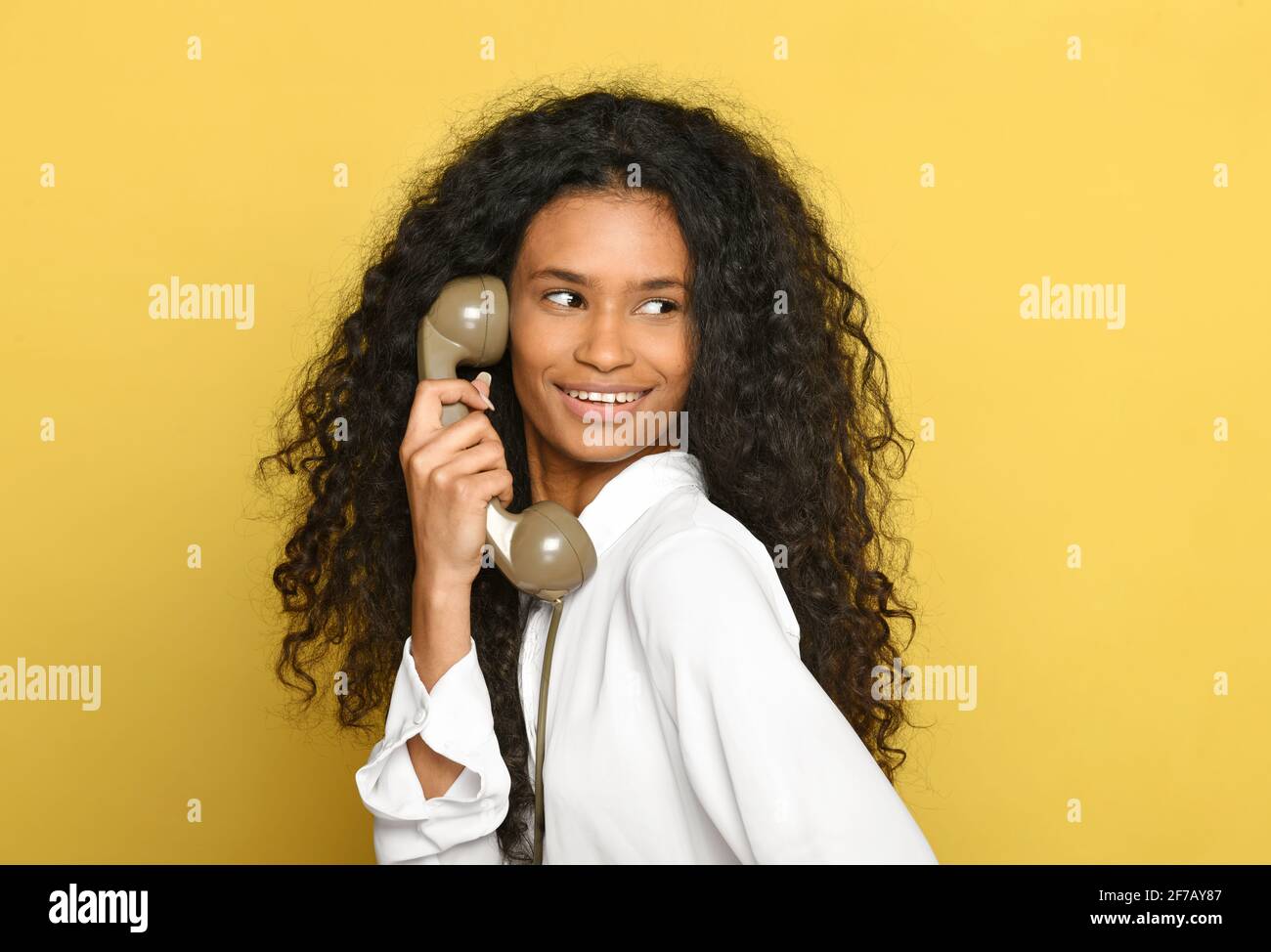 Happy smiling black woman holding a retro telephone receiver as she looks away over her shoulder on a yellow studio background in a close up head and Stock Photo