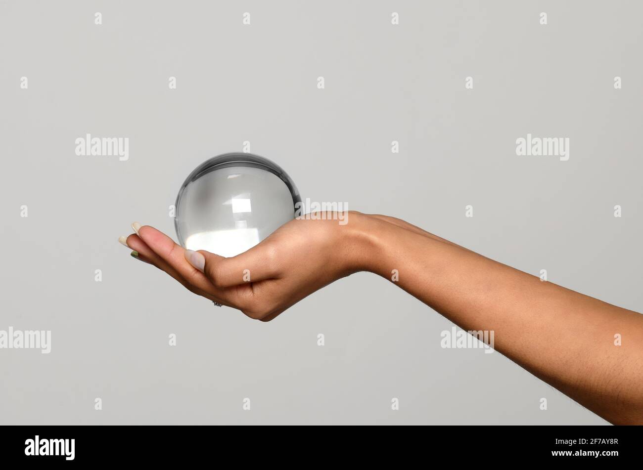Hand of a young black woman holding a transparent glass or crystal sphere, orb or ball cupped in her palm over a light grey studio background with cop Stock Photo