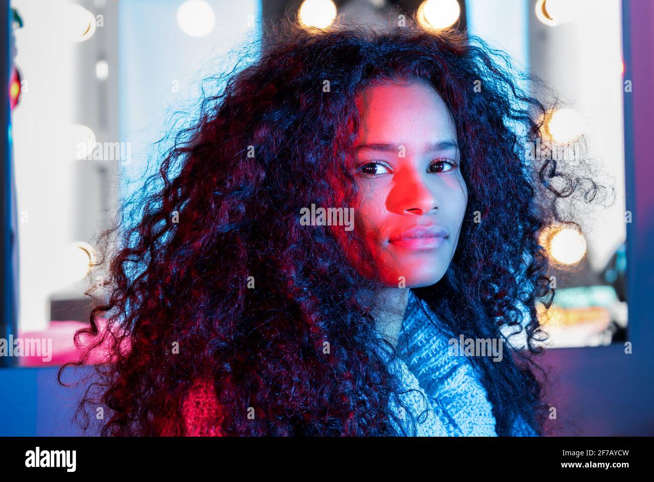 Cute attractive black woman frame by glowing lights with thick curly hair watching the camera with a sideways glance side lit by colorful pink studio Stock Photo