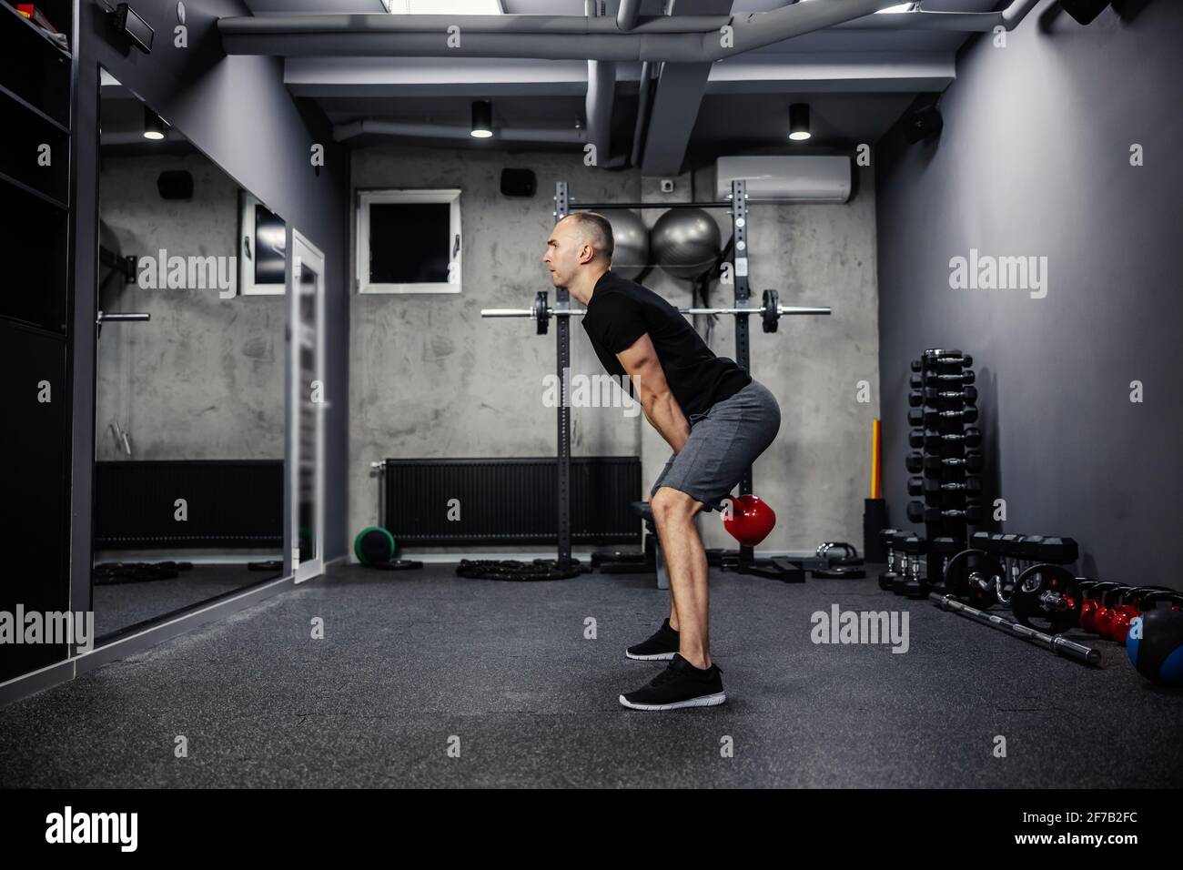 Dead lifting workout with a kettle bell. Man in sportswear with a black T-shirt stands with his legs spread and lifts a kettle bell with both arms in Stock Photo