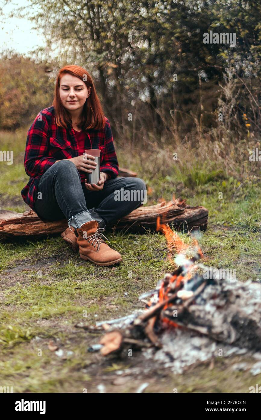 Woman traveler camping in the woods and resting by the fire after a hard day. girl holding hot tea or coffee in the thermos. Trekking, adventure and Stock Photo