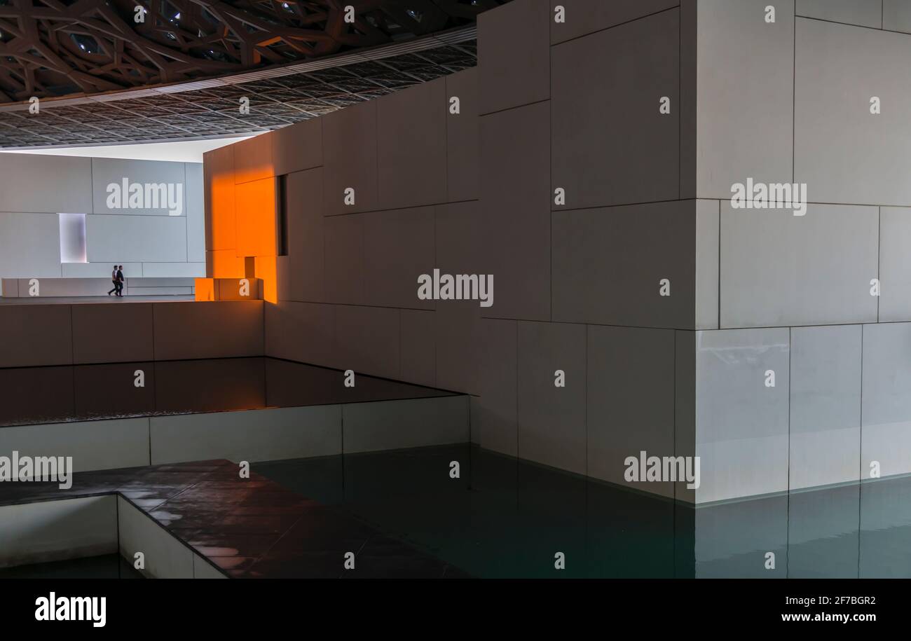 Two men walking in the direction of a white wall changed to orange due to the setting sun. Louvre museum. Abu Dhabi, United Arab Emirates. Stock Photo