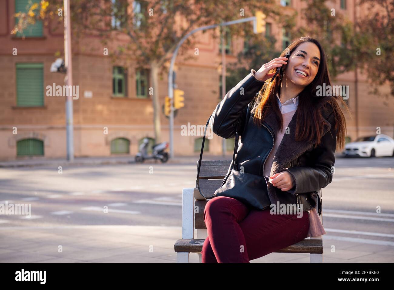 joyful young woman sitting on a street bench laughing while making a phone call, concept of communication and urban lifestyle, copyspace for text Stock Photo