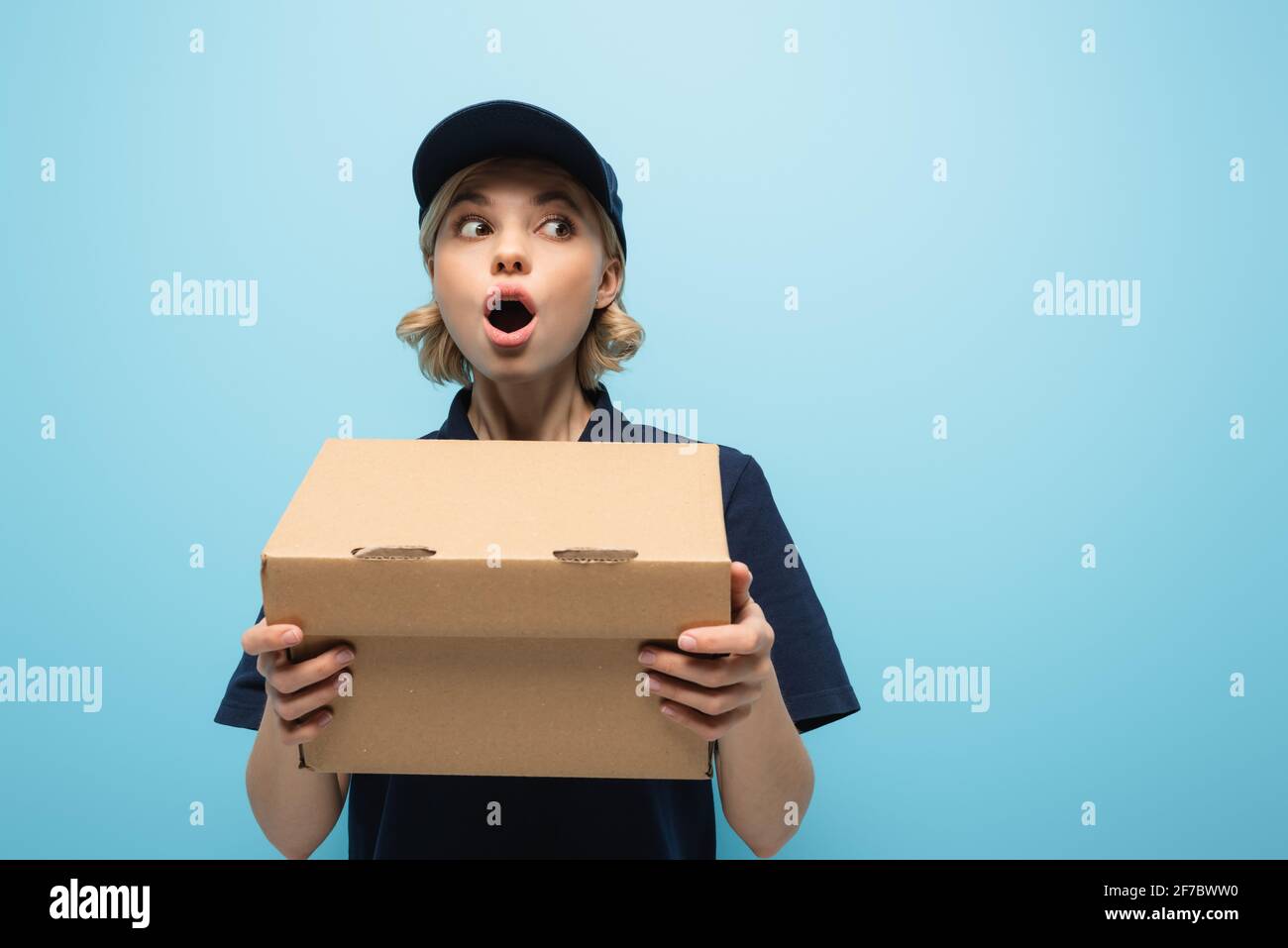 shocked courier looking away while holding parcel isolated on blue Stock Photo