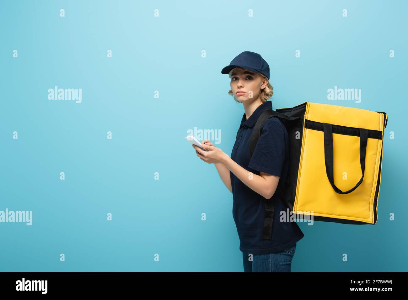 upset courier holding smartphone while carrying thermo bag on blue Stock Photo