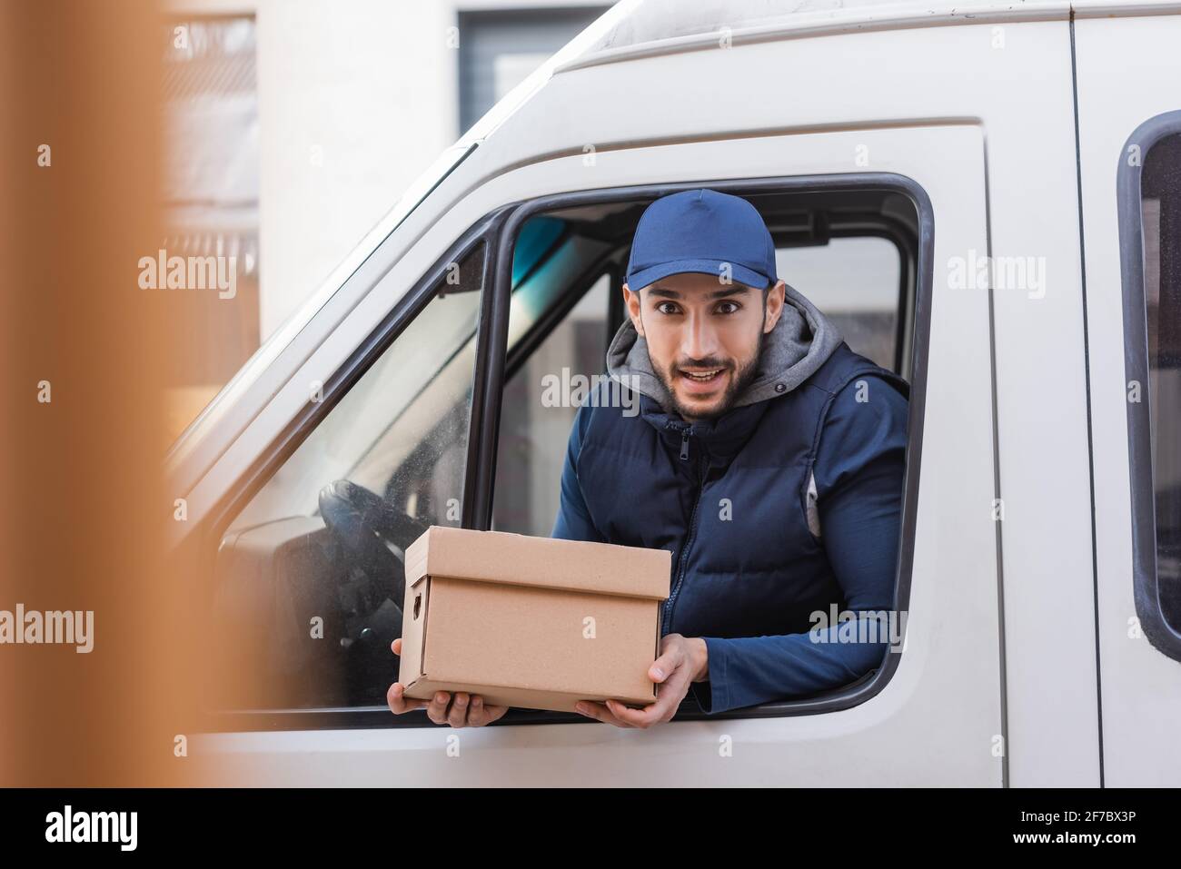 positive delivery man holding parcel while looking out car window on ...