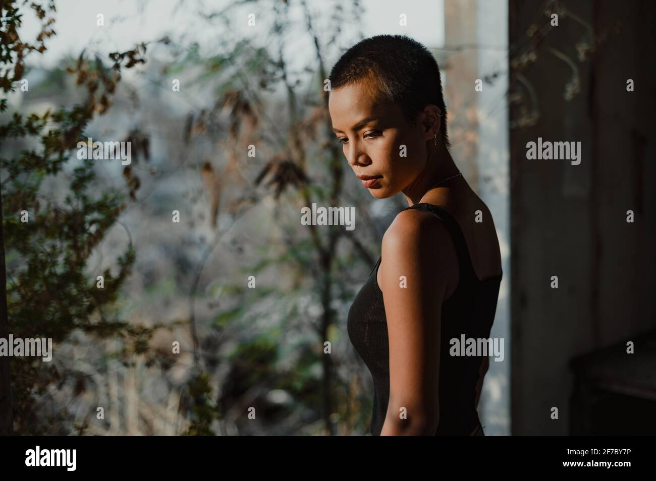 Portrait of a young short hair Asian woman standing by a big window looking down, with trees out of focus in the background. Stock Photo
