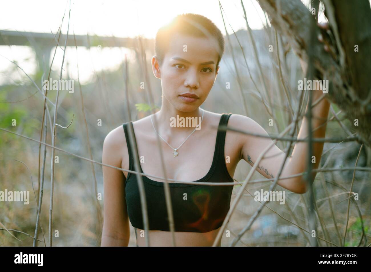 Closeup portrait of a young Asian ethnicity woman with short hair in nature, wearing a sports bra, looking at the camera. She is surrounded by bushes. Stock Photo