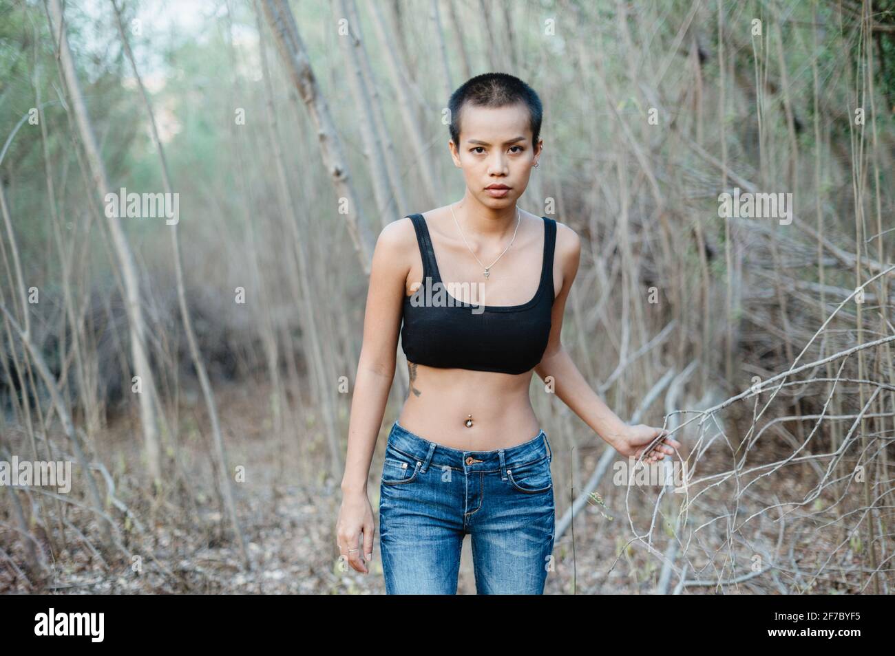 Portrait of a young Asian ethnicity woman with short hair in nature, wearing a sports bra, looking at the camera. She is surrounded by bushes. Stock Photo