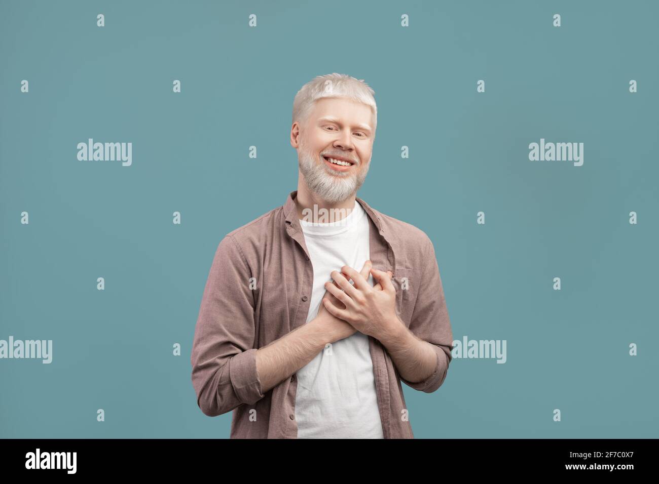 Sincere albino guy holding hands on chest, expressing gratitude and positive emotions on turquoise studio background Stock Photo